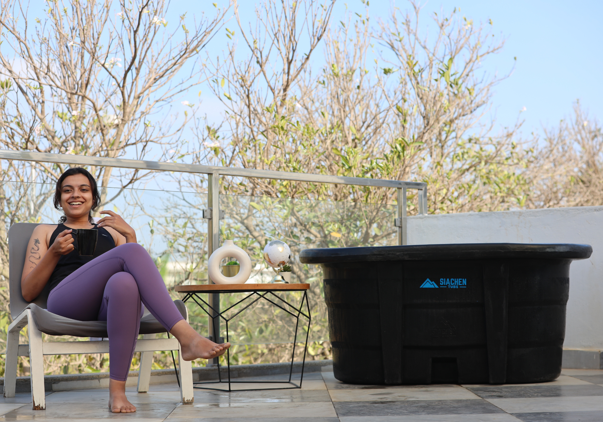 Woman sitting on a chair next to a ice bath hot tub on a patio with trees in the background