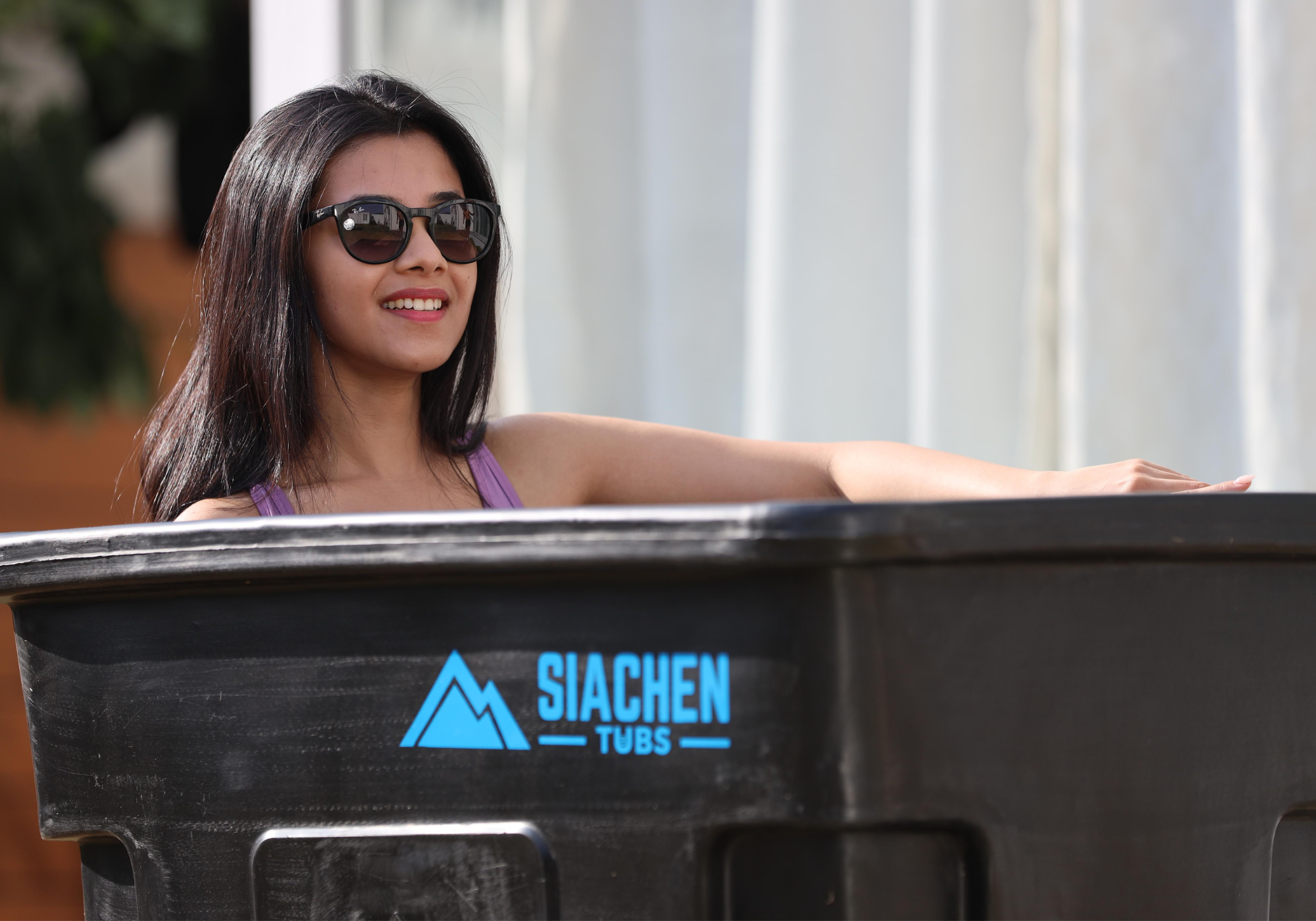 Woman sitting on a black ice bath tub with 'Siachen Tubs' logo, wearing sunglasses.
